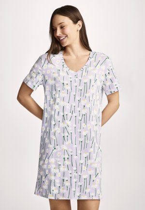 Jonge vrouw die lacht, gekleed in een lichtpaars jurkje met witte madeliefjes, staand met haar handen achter haar rug.