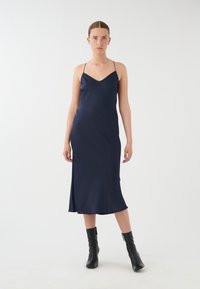 Robe nuisette satin marine avec fines bretelles, décolleté en V, silhouette ajustée et ourlet fluide. Portée avec des bottines noires à la cheville.