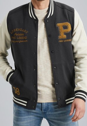 Mężczyzna noszący czarną i kremową kurtkę varsity z złotymi literami "Legendary Aviator PME Legend" i numerem "58", na beżowym swetrze i niebieskich dżinsach.