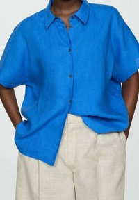 Camicia in lino blu con una vestibilità rilassata, maniche corte, chiusura a bottoni sul davanti e orlo leggermente curvato; abbinata a pantaloni beige chiaro.