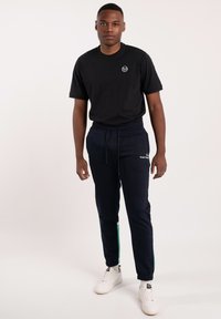 Svart t-shirt med en liten logotyp, marinblå mjukisbyxor med gröna detaljer, elastiska muddar och vita sneakers med en strukturerad yta.