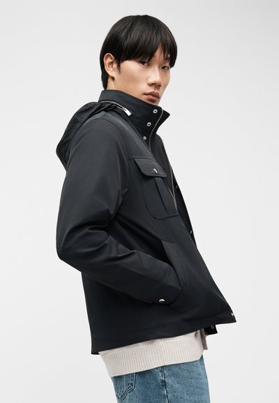 Νέος άνδρας που φοράει μαύρο μπουφάν με κουκούλα και μπροστινές τσέπες, ελαφρύ πουλόβερ και μπλε τζιν, κοιτάζοντας από πάνω του ώμου.