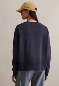 Granatowa bluza z prążkowanym obszyciem, luźnym krojem i wyraźną teksturowaną panelem na plecach; noszona z szarymi spodniami sportowymi i beżową czapką.