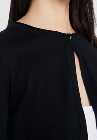 Schwarze gestrickte Strickjacke mit rundem Ausschnitt, ausgestattet mit einem einzigen Knopfverschluss. Der Stoff ist glatt und leicht strukturiert.