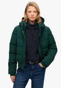Zielona pikowana kurtka z kapturem, zapinana na zamek i zatrzaski, noszona na ciemnoniebieskim swetrze z dzianiny i niebieskich dżinsach. Gładka faktura materiału.