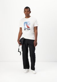 Camiseta blanca de algodón con gráfico de barco, combinada con pantalones cargo negros y zapatillas blancas. Pequeño bolso cruzado negro con asa.