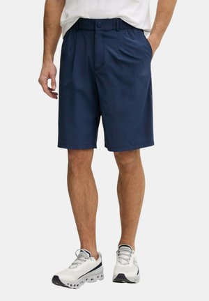 Uomo che indossa pantaloncini blu navy fino al ginocchio, scarpe da ginnastica bianche e camicia bianca, in piedi con una mano in tasca su sfondo bianco.