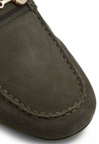 Gros plan sur une chaussure mocassin en daim vert olive avec coutures apparentes et détail en métal sur la bande élastique près du dessus.