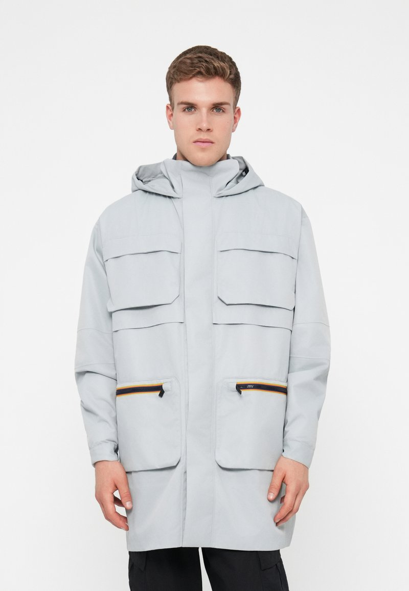 Heledad hallhallikaitsega heledat halli vihmamantlit, millel on kapuuts, kaks suurt rindu taskut ja kaks küljetaskut lukuga, millel on oranžid detailid.