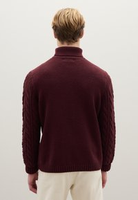 Burgunderfarbenes Strickpullover mit hohem Kragen und Zopfmuster-Ärmeln, mit gerippten Bündchen und Saum, zeigt eine strukturierte Oberfläche.