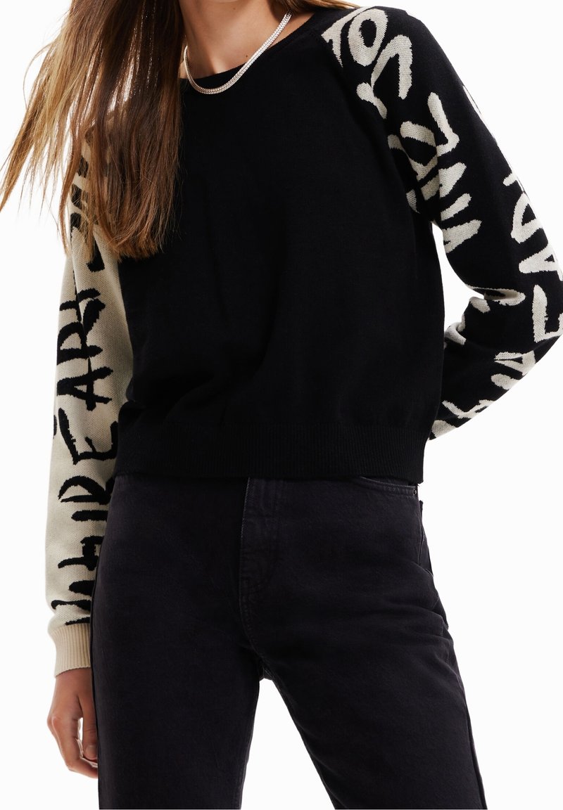 Femme portant un pull noir avec des manches blanches contrastantes comportant un texte noir en gras et un pantalon sombre, coupé au milieu du torse.