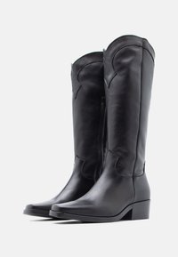 Čierne kožené čižmy po kolená s mierne zaobleným horným okrajom, bočným zipsom, nízkym hrubým podpätkom a dekoratívnym prešívaním pozdĺž švov.