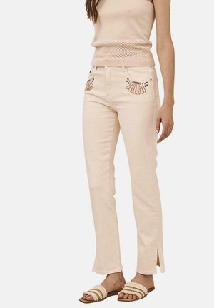 Vaqueros de denim beige con dobladillos abiertos, que presentan coloridos patrones de conchas bordados en los bolsillos delanteros. Llevados con una blusa beige a juego.