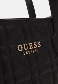 Sac à main en cuir matelassé noir avec logo doré "GUESS EST 1981" et deux poignées visibles en haut.