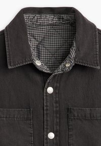 Černá džínová košile s prošitou šedou a černou kostkovanou podšívkou, límečkem do špičky, stříbrnými patentními knoflíky a přední kapsou na prsou.