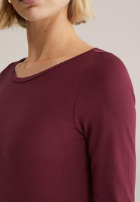 Bordowa bluzka z długim rękawem o okrągłym dekolcie i dopasowanym kroju, wykonana z gładkiej, elastycznej tkaniny. Widoczne przeszycia na ramieniu.