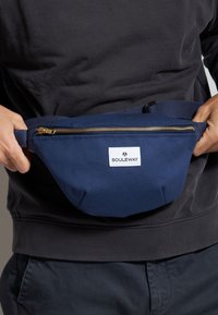 Riñonera de tela azul marino con una cremallera dorada, correa ajustable y una etiqueta blanca de marca. Presenta una forma redondeada y un diseño duradero.