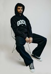 Schwarzer Hoodie mit "GOOD"-Text, lässig geschnittene Hose und schwarze Sneakers. Stricktextur beim Sweater, glattes Material bei der Hose und klassisches Schnürdesign bei den Schuhen.