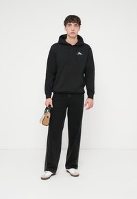 Sweat à capuche noir avec poche avant et cordons de serrage, associé à un jean noir à jambes larges. Tient un petit sac à main beige. Des baskets blanches complètent le look.