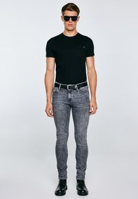 Mees, kes kannab musti päikeseprille, musta liibuva T-särgi, hallid kitsad teksad musta vööga ja mustad saapad, seisab ühevärvilise tausta ees.