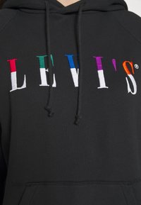 Sweat à capuche noir avec des lettres multicolores cousues formant "LEVI'S" sur la poitrine, et des cordons noirs suspendus à la capuche.