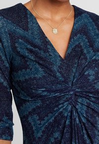 Laivastonsininen mekko, jossa on teksturoidusta kankaasta tehty kiiltävä kuviointi. Solmisolmu vyötäröllä ja V-pääntie. Kultainen riipuskaulakoru.