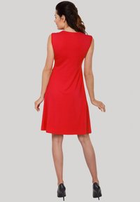 Rotes ärmelloses Kleid mit einem schwingenden Rock, aus glattem Stoff. Es hat einen bescheidenen Ausschnitt und wird mit schwarzen High Heels kombiniert.