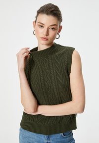 Suéter de punto sin mangas de color verde oliva con patrón de trenzas, cuello y dobladillo acanalados, combinado con jeans de mezclilla azules, manos descansando sobre el brazo.