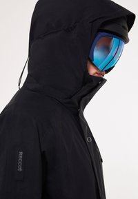 Veste noire imperméable avec capuche, dotée d'une fermeture éclair brillante et de la marque RECCO sur la manche, portée avec des lunettes de protection miroitées bleues.