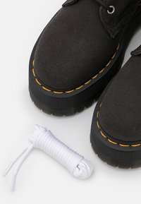 Czarne płócienne buty na grubej czarnej podeszwie, z teksturowanym wykończeniem, z żółtymi szwami. Obok znajduje się sprężyna białych sznurowadeł.