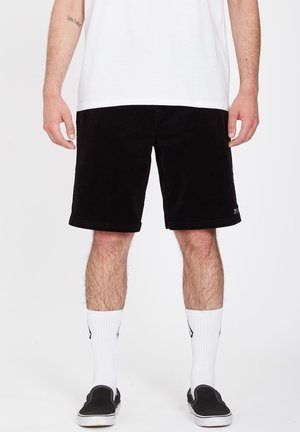 Sorte fløjlsshorts med en afslappet pasform, der har forlommer og en kant, der sidder lige over knæet. Båret med hvide crew-sokker og sorte slip-on sko.