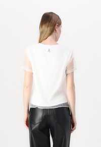 T-shirt bianco con maniche corte trasparenti, collo rotondo e una texture di tessuto leggero. Presenta un piccolo logo argentato sul retro.