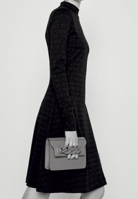 Robe noire à manches longues avec un motif texturé de texte. Présente une silhouette ajustée, un col montant, et un sac à main gris contemporain avec un accent en chaîne.