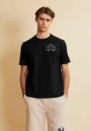 Giovane uomo con capelli castani corti che indossa una maglietta nera e pantaloni beige chiaro, entrambi con piccoli giocatori di polo ricamati.