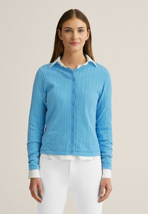 Ung kvinde iført en lyseblå tekstureret cardigan med knapper over en hvid skjorte med krave og hvide bukser, stående foran en ensfarvet baggrund.