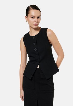 Donna con capelli tirati indietro che indossa un gilet nero senza maniche con bottoni e una gonna nera abbinata su uno sfondo chiaro e semplice.