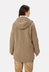 Giubbotto marrone con cappuccio e rivestimento in pelliccia sintetica, dallo stile comodo con maniche elastiche, mostrato da dietro, abbinato a pantaloni chiari.