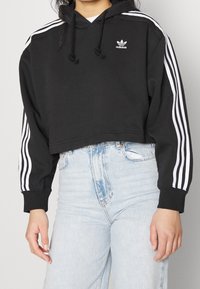 Felpa nera corta con logo adidas bianco, caratterizzata da tre strisce bianche lungo le maniche. Abbinata a jeans azzurri a vita alta.