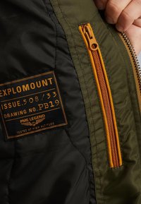Intérieur d'une veste isolée vert olive et noire avec une poche zippée orange et une étiquette présentant "EXPLOROMOUNT" et des détails de logo.