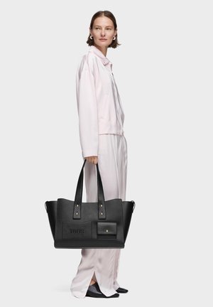 Donna con abito di seta rosa pallido, che tiene una grande borsa tote in pelle nera con tasche frontali e manici corti.