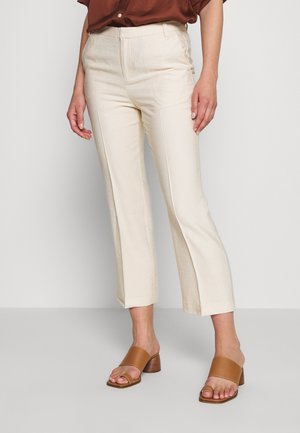 Kvinde iført cremenfarvede bukser med lige ben og forreste syninger samt tanfarvede blokhæl-sandaler med åbne tæer, stående foran en ensfarvet baggrund.