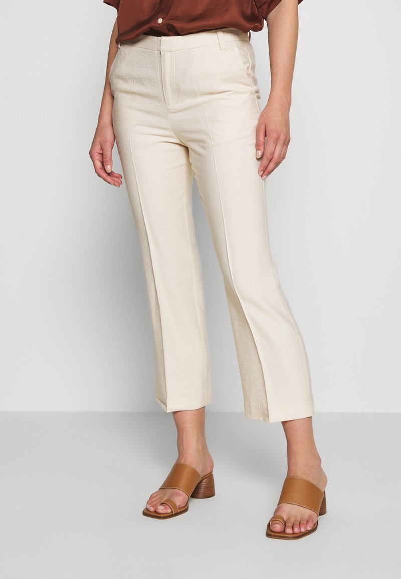 Femme portant un pantalon droit crème avec des coutures à l'avant et des sandales à talons blocs ouvertes couleur beige, debout devant un fond uni.