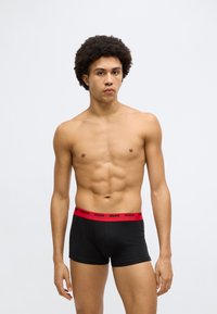 Cuecas boxer pretas com uma cintura elástica vermelha com a marca "HUGO". O tecido parece macio, e o ajuste é justo e contemporâneo.