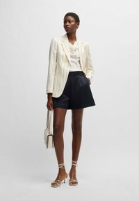 Blazer mit weißen und schwarzen Nadelstreifen, kombiniert mit einem beigen Oberteil und marineblauen Shorts. Die Accessoires umfassen eine hellfarbige Handtasche und Riemchensandalen.