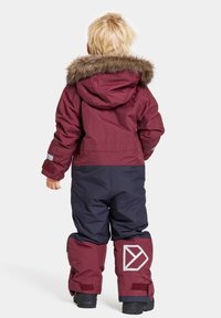 Barnens vinteroverall, vinröd överdel med konstfjälls huva, marinblå byxor, reflektorer och stor logotyp på vänster ben. Väderbeständigt tyg.