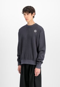 Giovane uomo con capelli corti e scuri che indossa una camicia a maniche lunghe grigio scuro con texture e pantaloni neri larghi su uno sfondo bianco.
