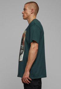 Teal bomulds T-shirt med en løs pasform, der har et grafisk motiv på fronten. Designet inkluderer trykt tekst og illustrationer, samt en rund halsudskæring.