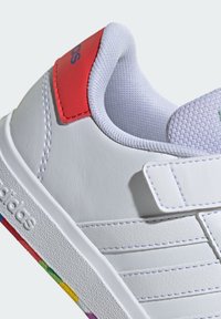 Sneaker en cuir blanc avec un onglet de talon rouge, semelle en caoutchouc texturée et détail de bande multicolore à la base. Comprend une sangle à crochet et boucle.