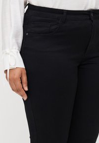 Black denim jeans with a slim fit, featuring a pocket on the left side and a smooth texture. Paired with a white shirt with tied sleeves.