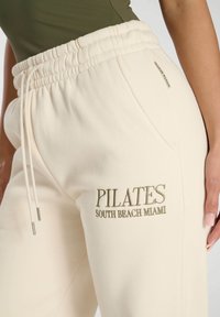 Pantalones de chándal color crema con una cinturilla elástica, que presentan bordados de "PILATES" y "SOUTH BEACH MIAMI" en dorado. Textura suave, estilo casual.